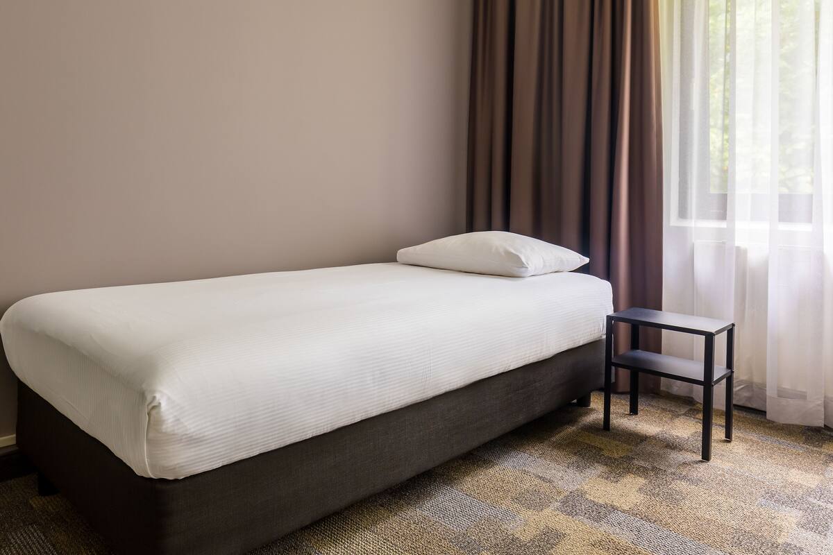 A single bed with a white mattress and pillow, a small bedside table, and a window with curtains.