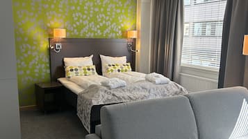 Een hotelkamer met een bed, een grijze fauteuil, een nachtkastje en een raam met gordijnen.