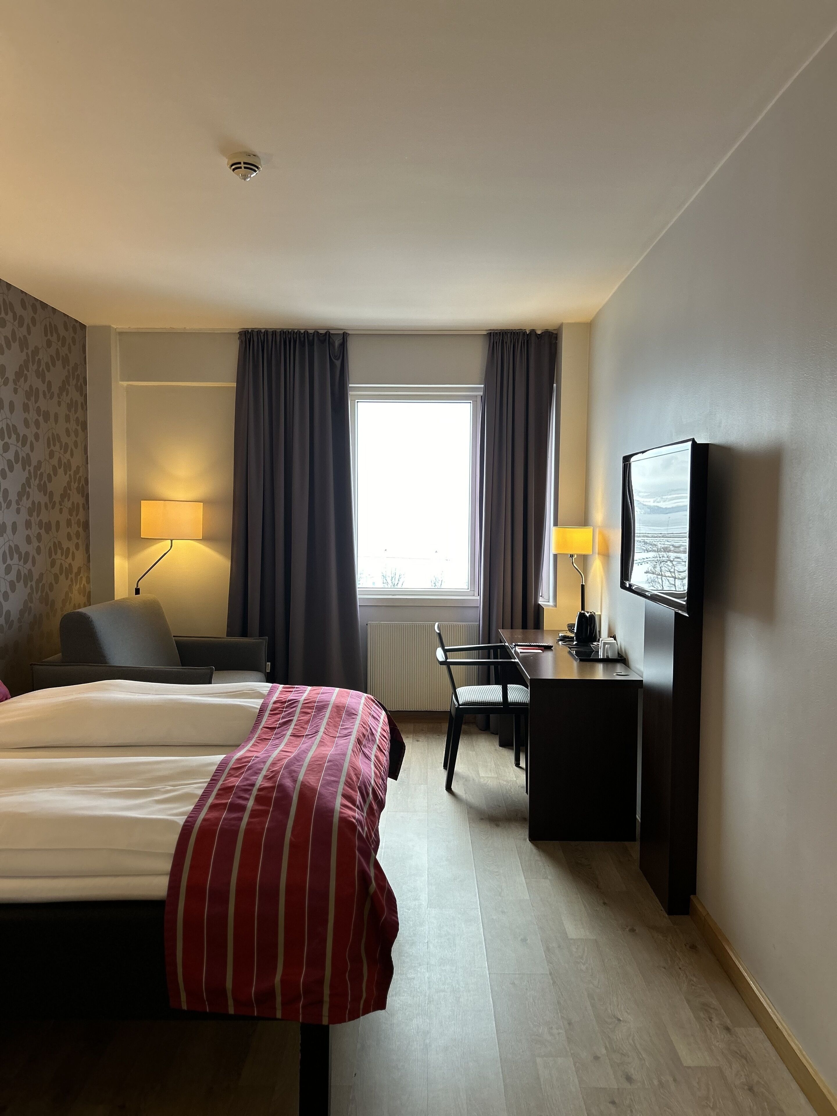 A hotel room with a bed, a desk, a chair, a TV, and a window with curtains.