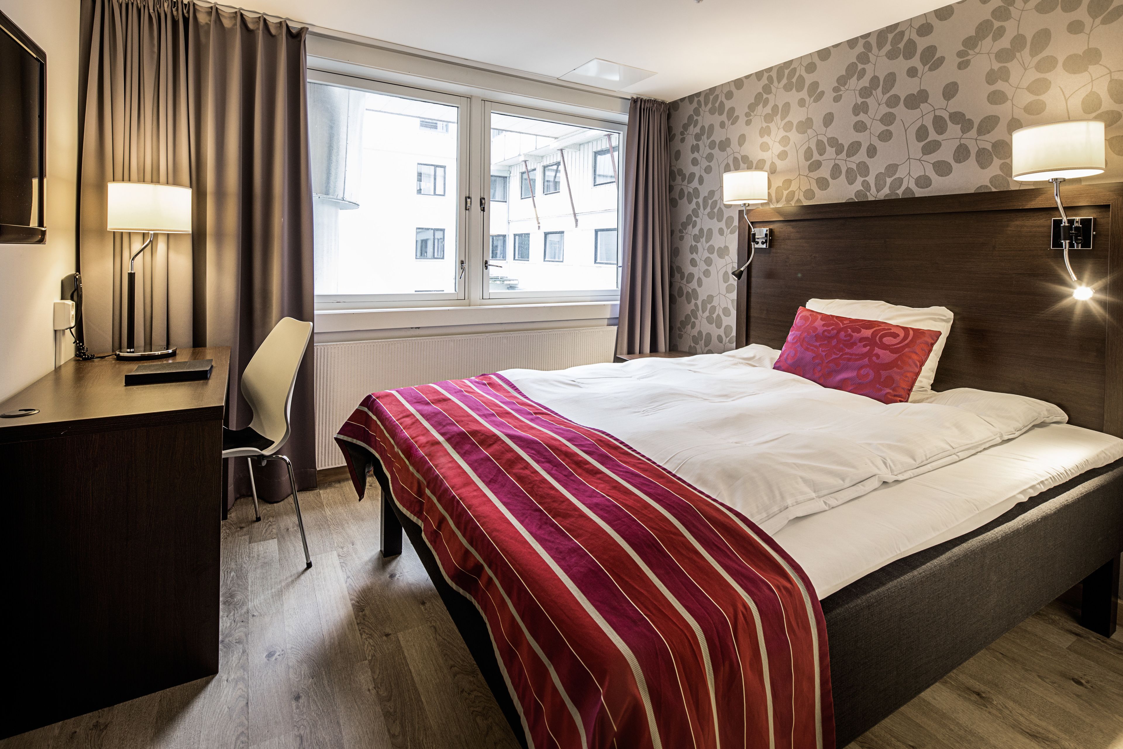A hotel room with a large bed, a desk, a chair, a window with curtains, and a wall with a leaf pattern.