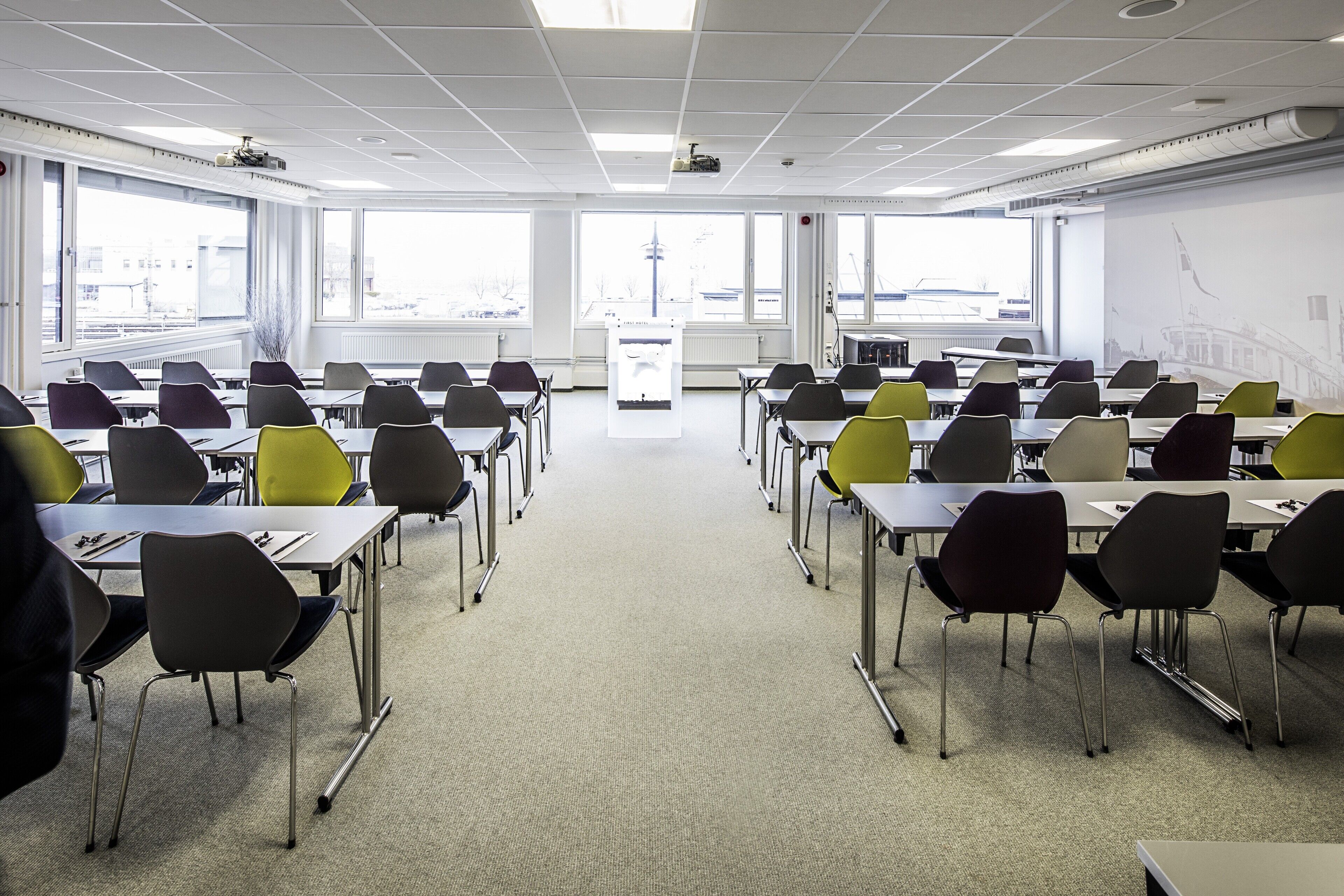 Une salle de conférence avec des rangées de tables et de chaises, de grandes fenêtres et un écran de projection.