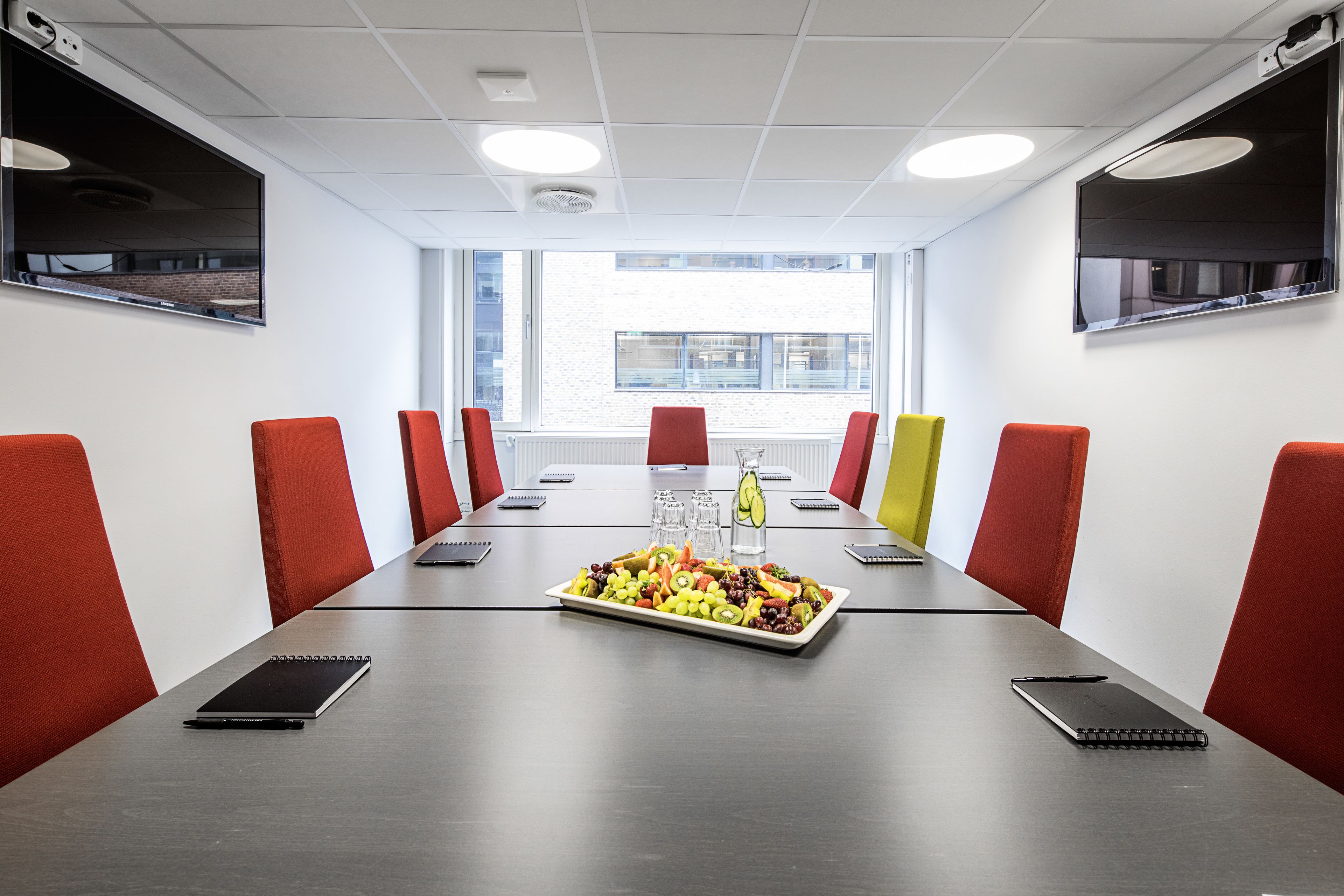 Ein Konferenzraum mit einem langen Tisch, roten und gelben Stühlen, einer Obstplatte und einem Laptop auf dem Tisch.