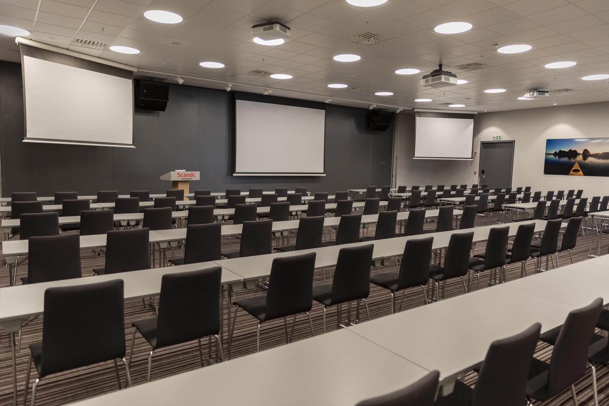 Un salón de conferencias con filas de mesas y sillas, una pantalla de proyector y una pizarra grande.