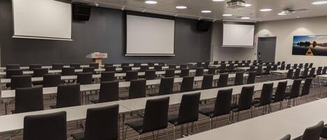 Un salón de conferencias con filas de mesas y sillas, una pantalla de proyector y una pizarra grande.