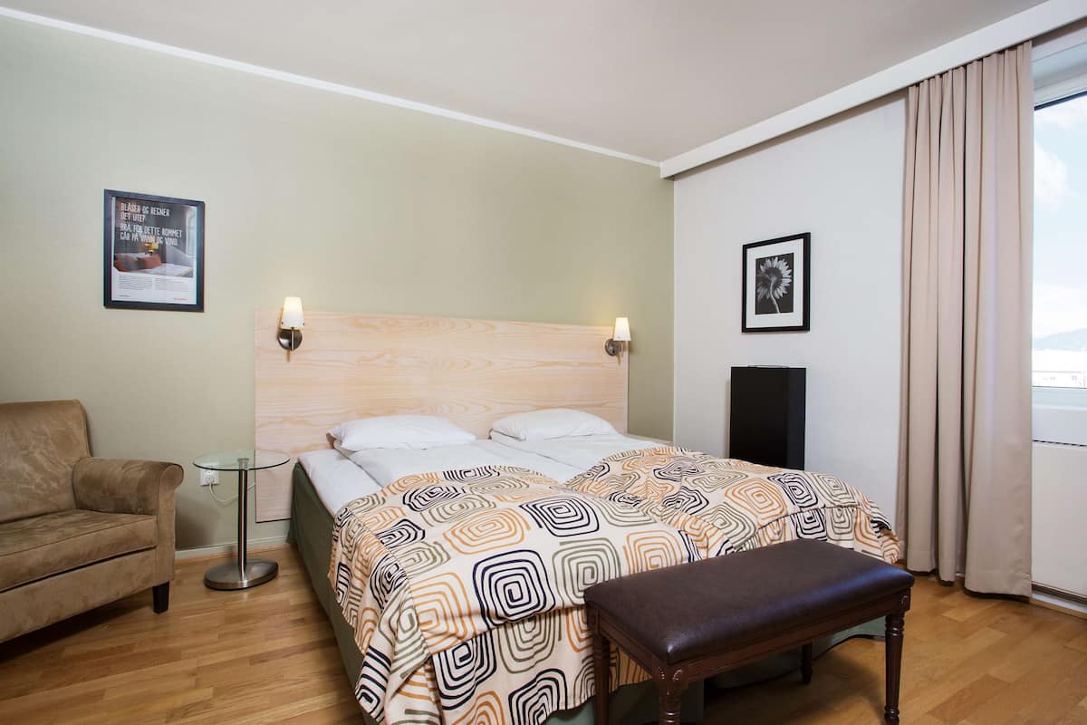A hotel room with a wooden headboard, a bed with patterned bedding, a bench, a small round table, a sofa, and a framed picture on the wall.