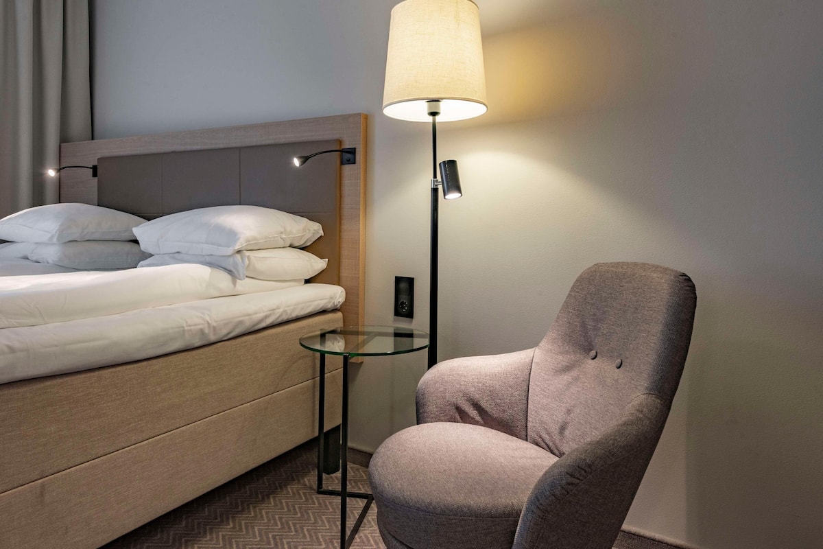 A hotel room with a bed, a chair, a lamp, and a small table.