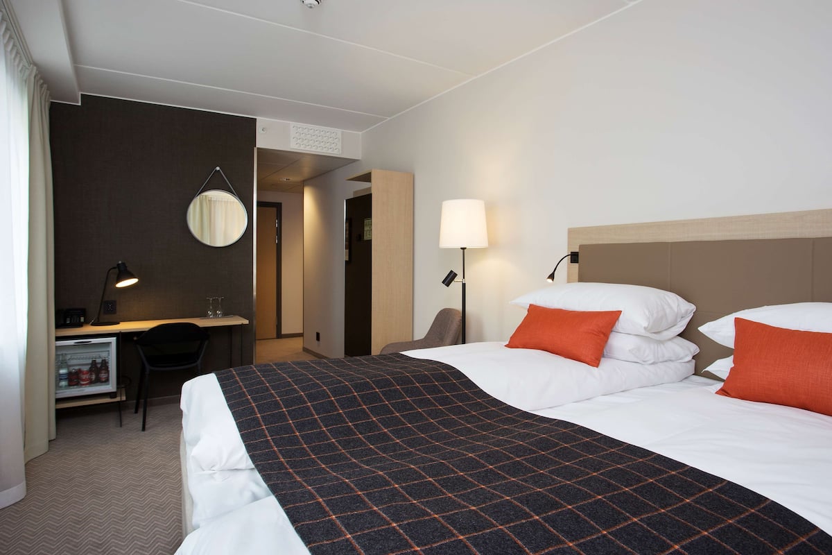 A hotel room with a bed, a desk, a chair, a lamp, and a refrigerator.