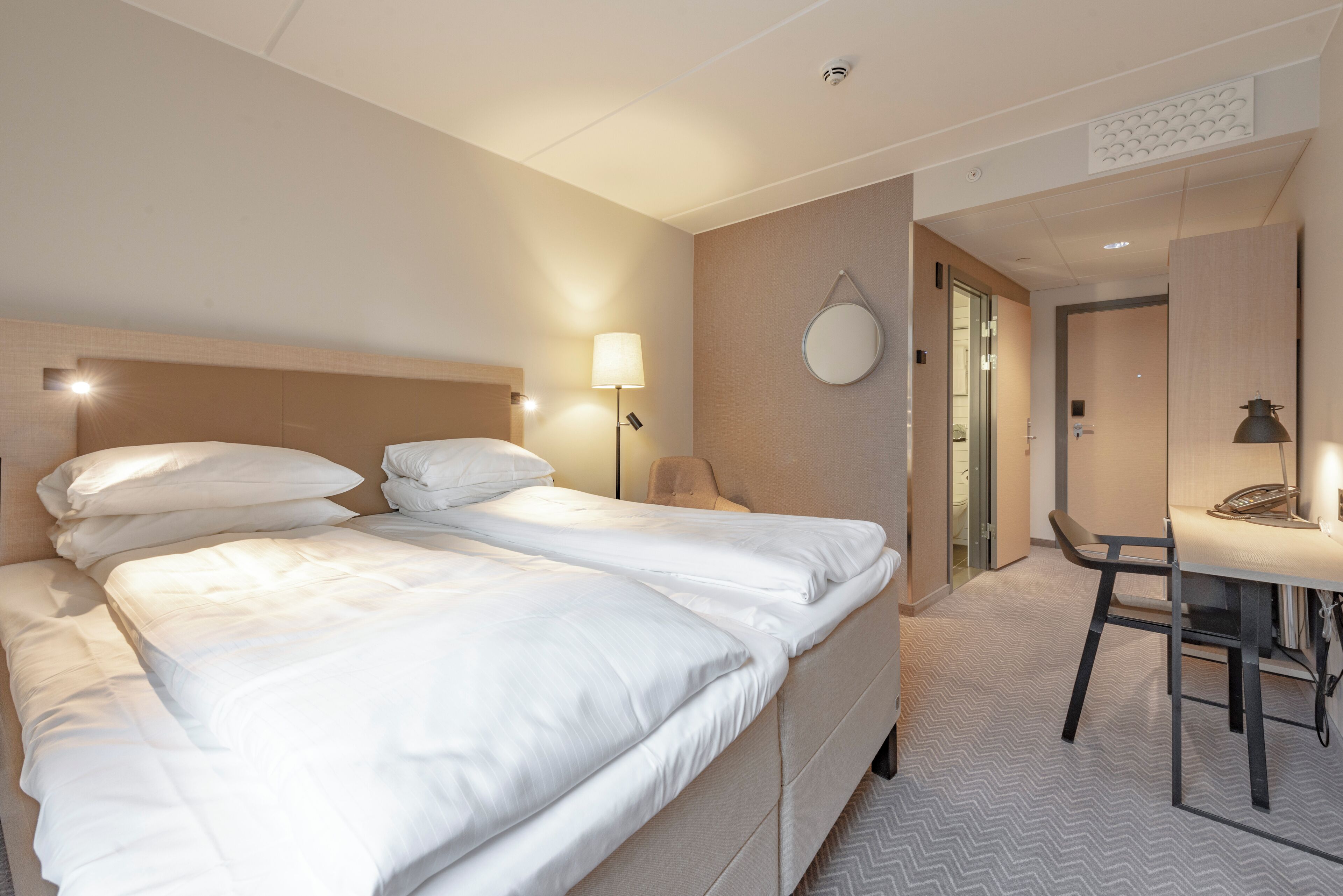 A hotel room with a double bed, bedside lamps, a round mirror, a desk with a telephone, and a chair.