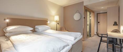 A hotel room with a double bed, bedside lamps, a round mirror, a desk with a telephone, and a chair.