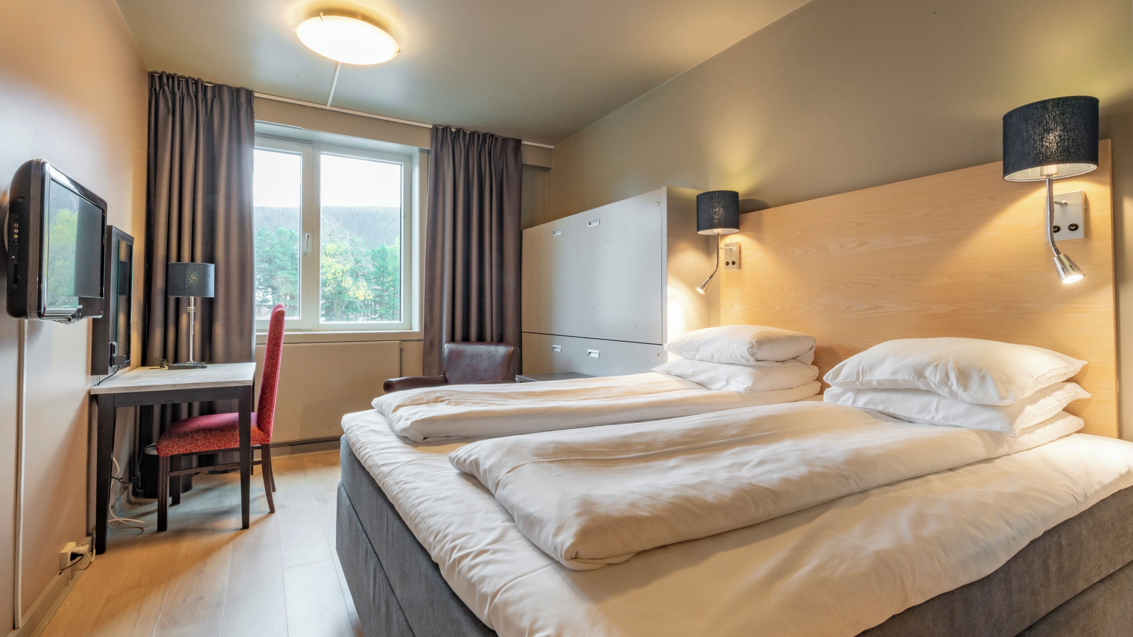 A hotel room with two beds, a desk, a chair, a TV, and a window with curtains.