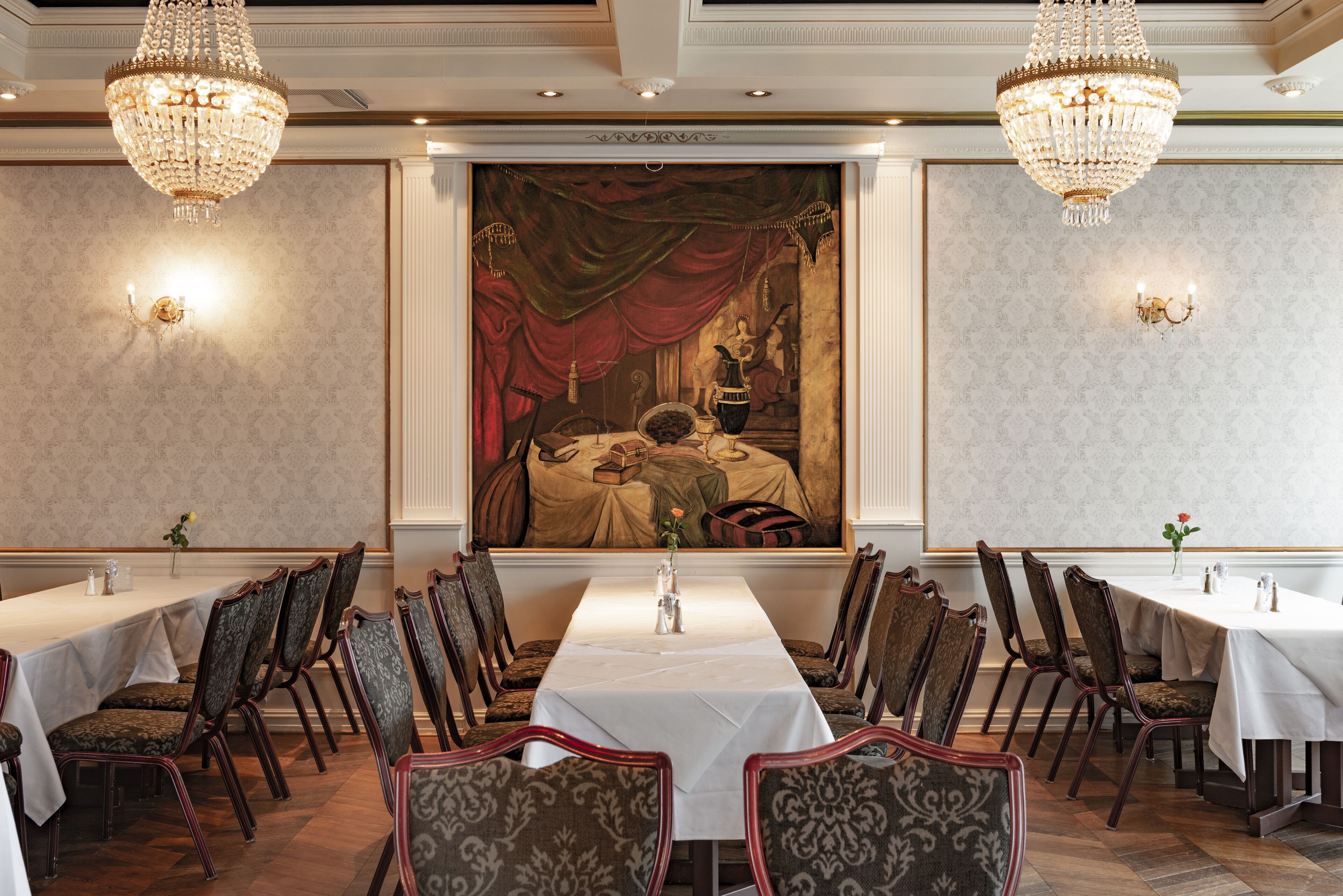 A formal dining room with a long table, chairs, chandeliers, and a large painting on the wall.