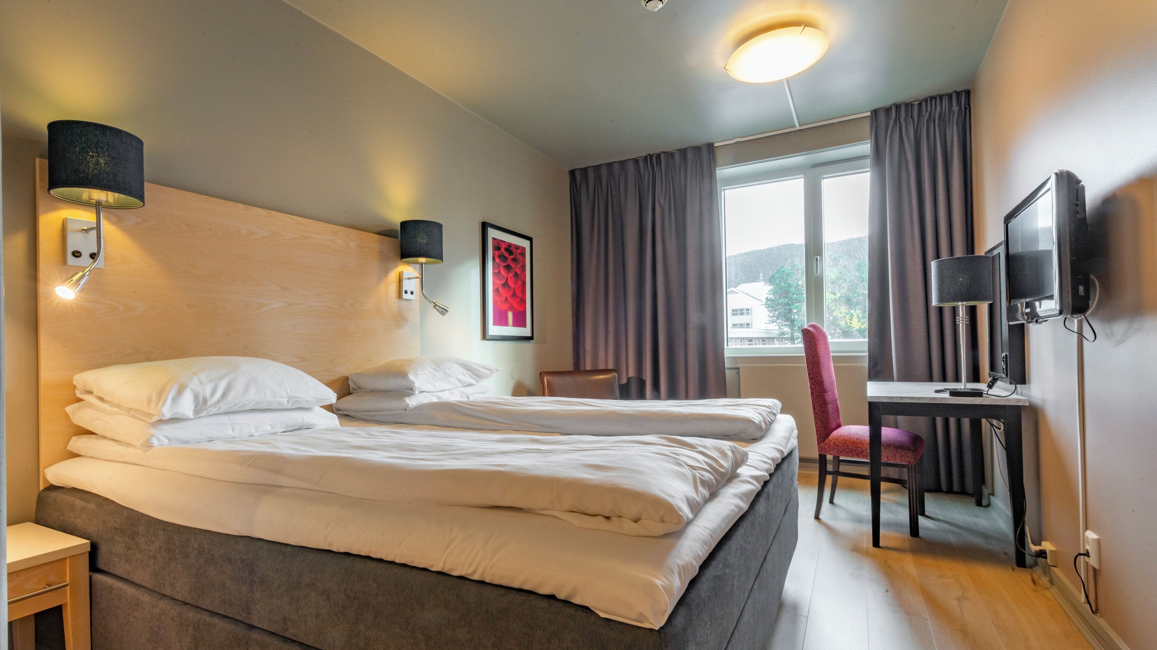 A hotel room with a bed, bedside lamps, a desk with a chair, a television, and a window with curtains.
