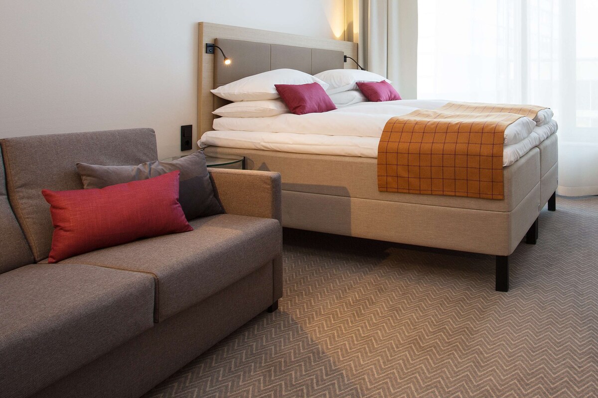 Una habitación de hotel con sofá, cama con manta de cuadros y mesita de noche.