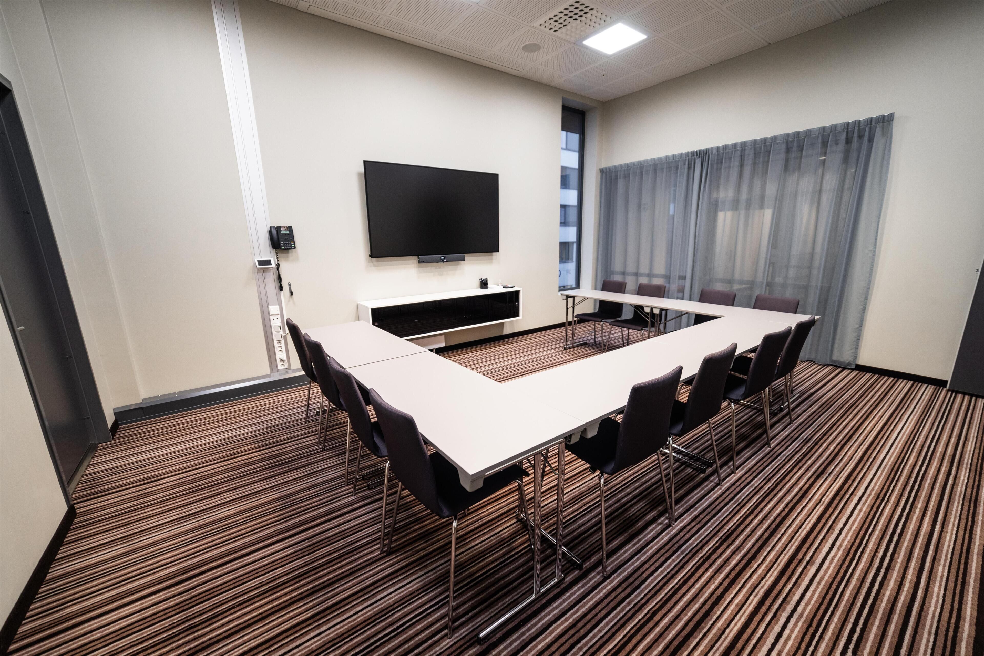 A conference room with a long table, chairs, a flat-screen TV, and a window with curtains.