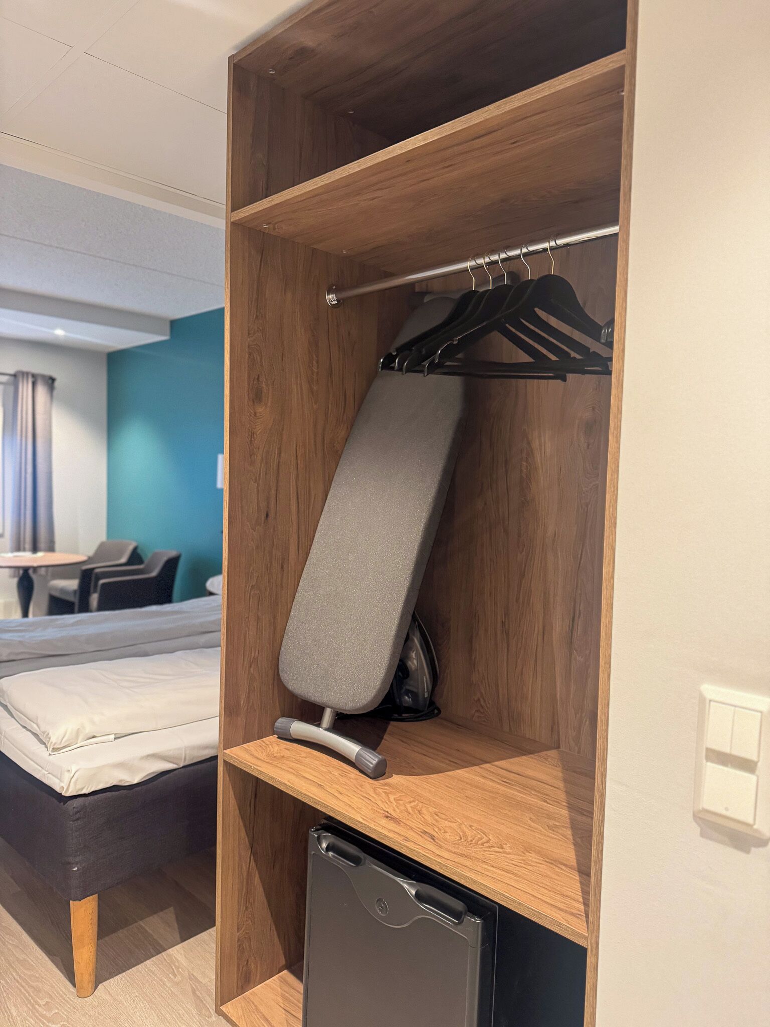 Ein kompaktes Hotelzimmer mit Bett, Schreibtisch mit Stuhl, einer kleinen Küchenzeile und einem Holzschrank mit Kleiderhaken.