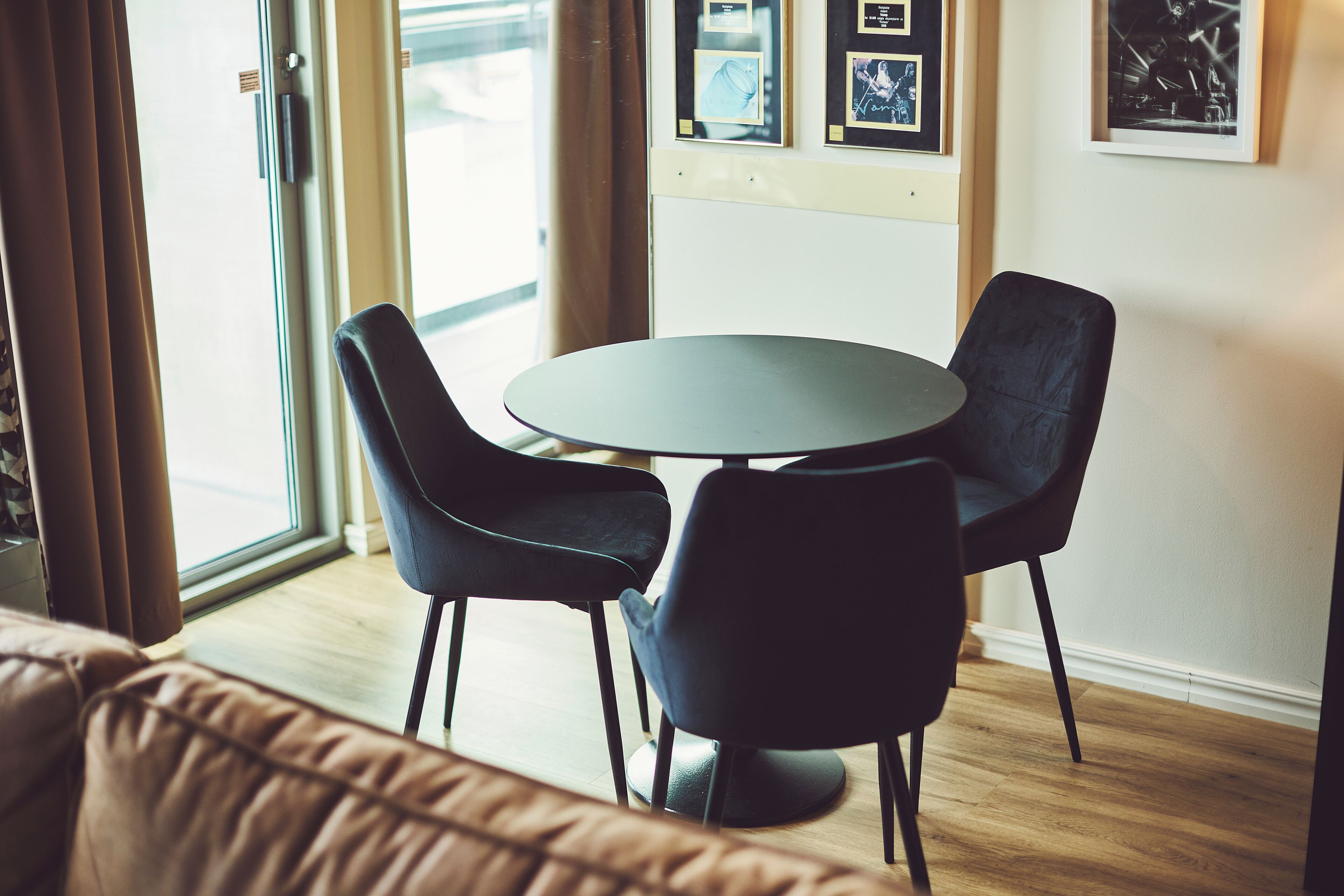 A modern room with a round table and four chairs, large windows with curtains, and framed pictures on the wall.