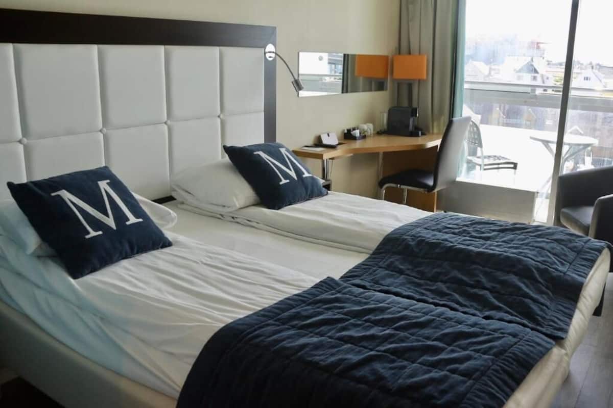 Hotellihuone, jossa on sänky, kaksi suurikokoista koristetyynyä, työpöytä, tuoli ja näkymä rakennuksiin.