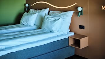 Ein ordentlich bezogenes Bett mit weißen Leinen und Kissen, ein Kopfteil mit einer Lampe sowie ein kleiner Nachttisch mit einem Lautsprecher.