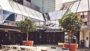 An outdoor seating area with tables and chairs, two large potted trees, and a building with a glass facade and a sign reading "Quality Hotel Maritim".