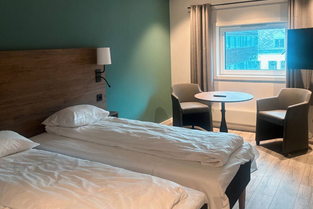 Hotellihuone, jossa on suuri sänky, kaksi tuolia, pieni pöytä ja ikkuna, jossa on verhot.