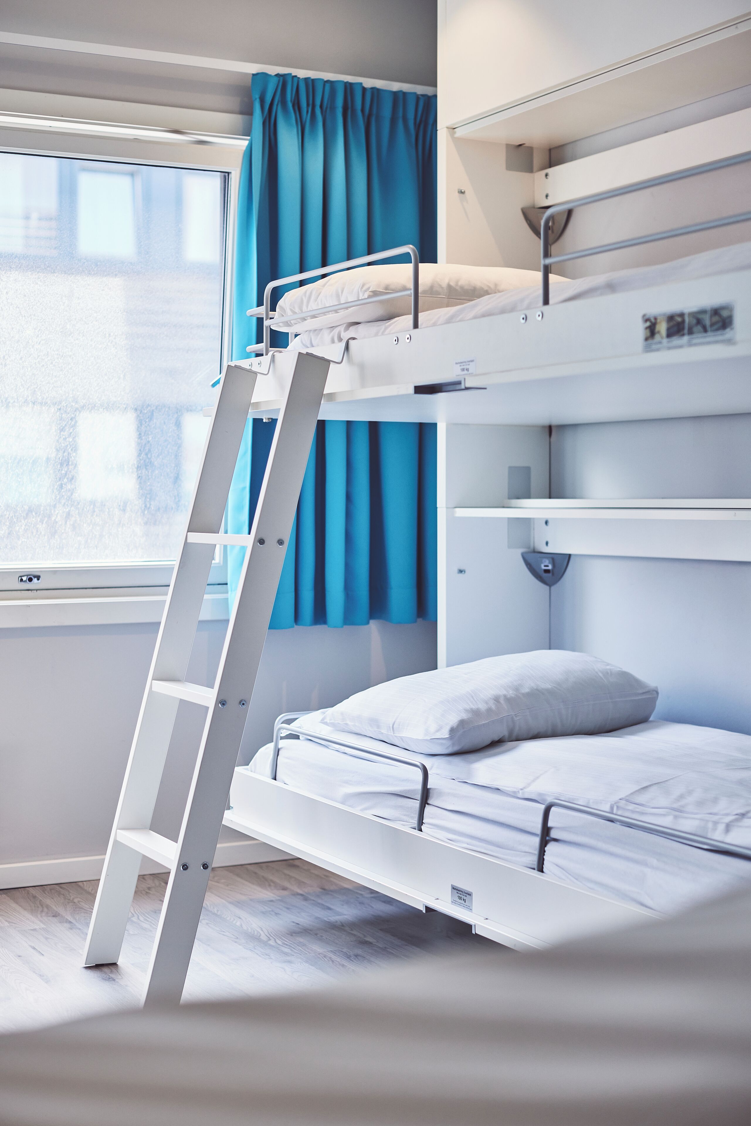 A bunk bed with a ladder, a window with blue curtains, and a white wall.