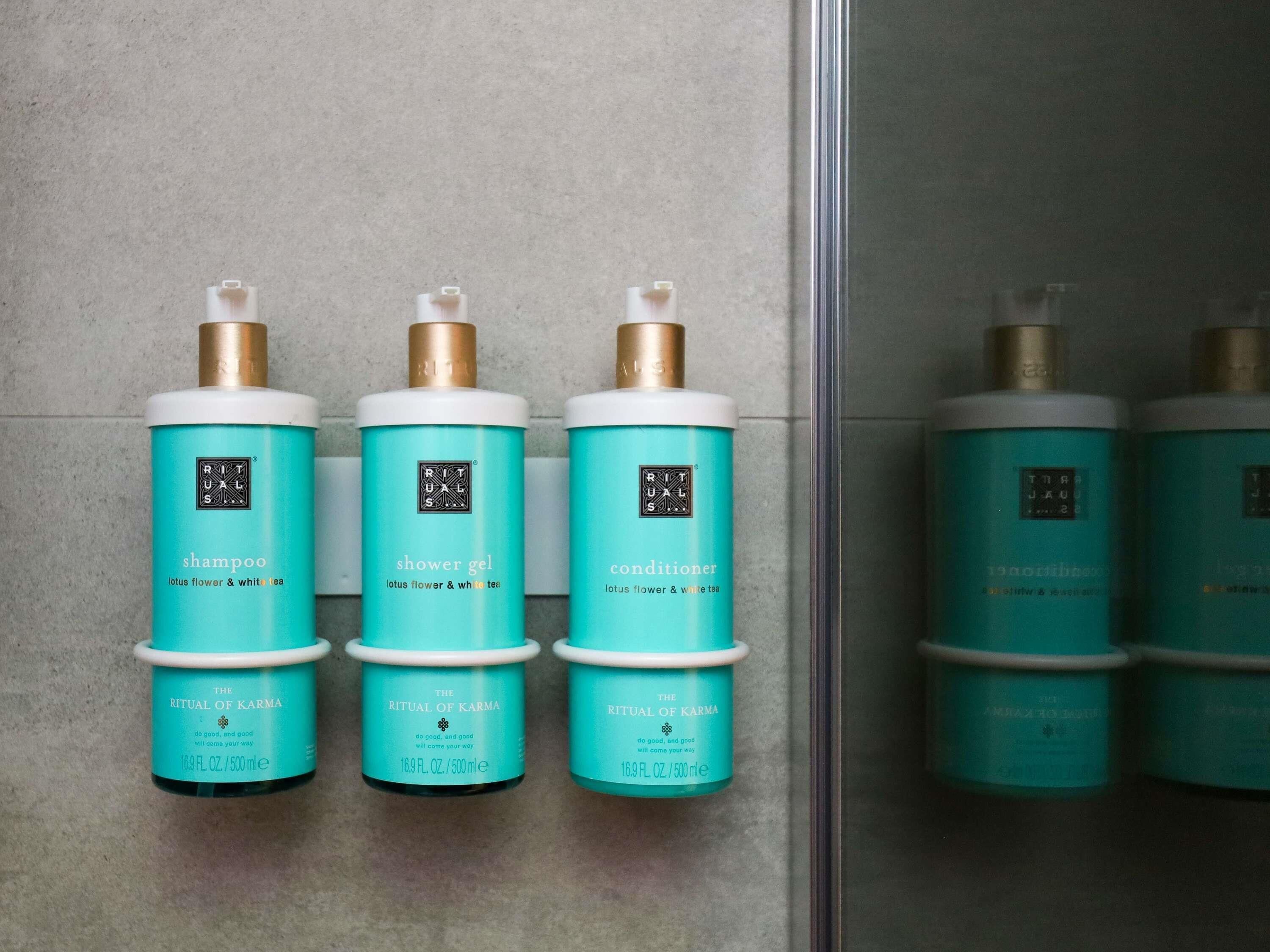Tres botellas de productos 'Ritual de Karma': champú, gel de ducha y acondicionador, dispuestas sobre una pared de azulejos.