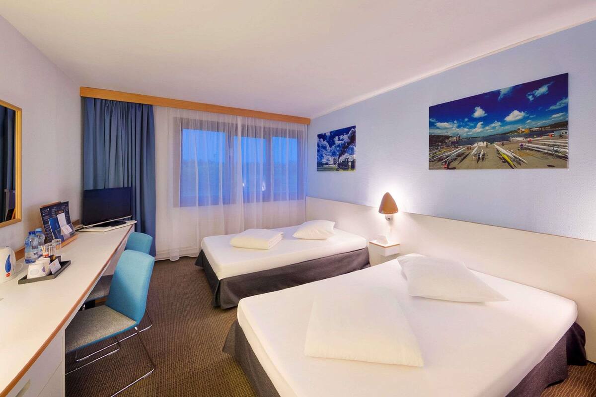A hotel room with two beds, a desk, a chair, a TV, and two paintings on the wall.