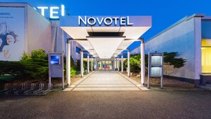 The exterior of a Novotel hotel with a modern design, featuring a prominent entrance, a digital information board, and a clear pathway leading to the entrance.