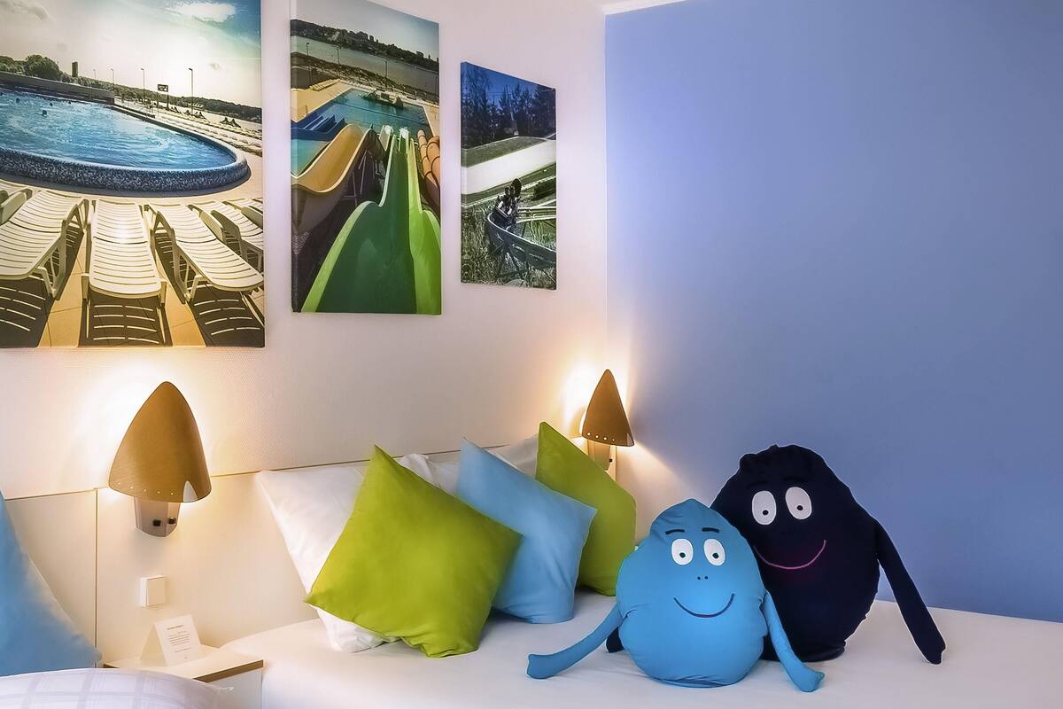 A hotel room with a bed, pillows, a nightstand, a lamp, and wall art depicting a pool and slides.