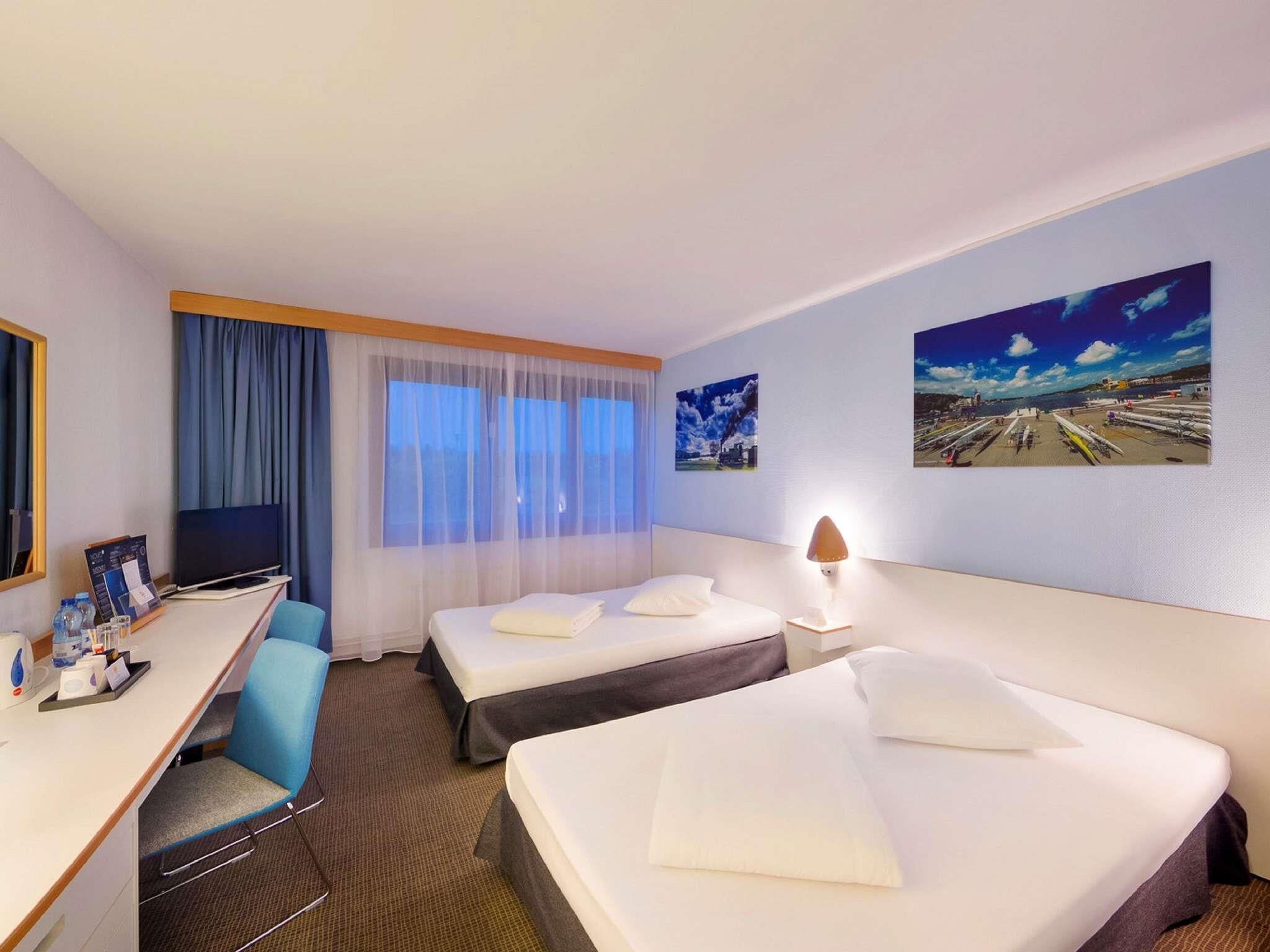 Ein Hotelzimmer mit zwei Betten, einem Schreibtisch, einem Sessel, einem Fernseher und zwei Wandgemälden.