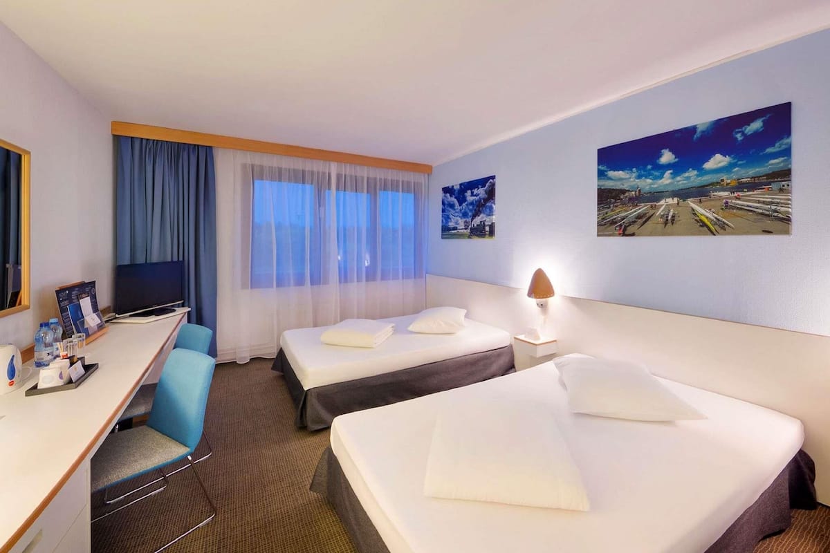 Ein Hotelzimmer mit zwei Betten, einem Schreibtisch, einem Sessel, einem Fernseher und zwei Wandgemälden.