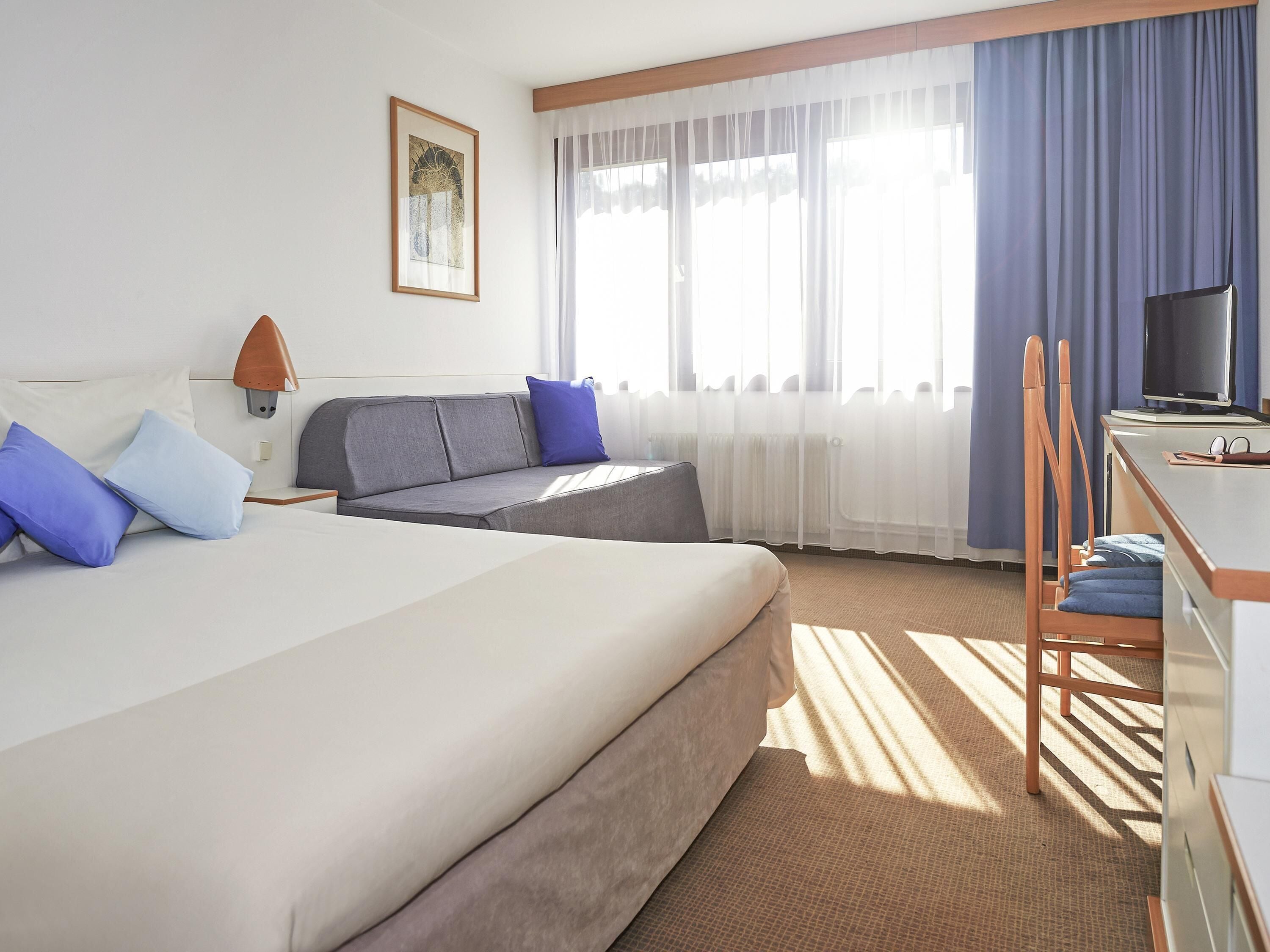 Habitación de hotel con cama, sofá, escritorio, silla, televisor y ventana con cortinas.
