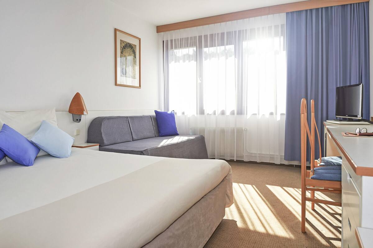 A hotel room with a bed, a sofa, a desk, a chair, a television, and a window with curtains.