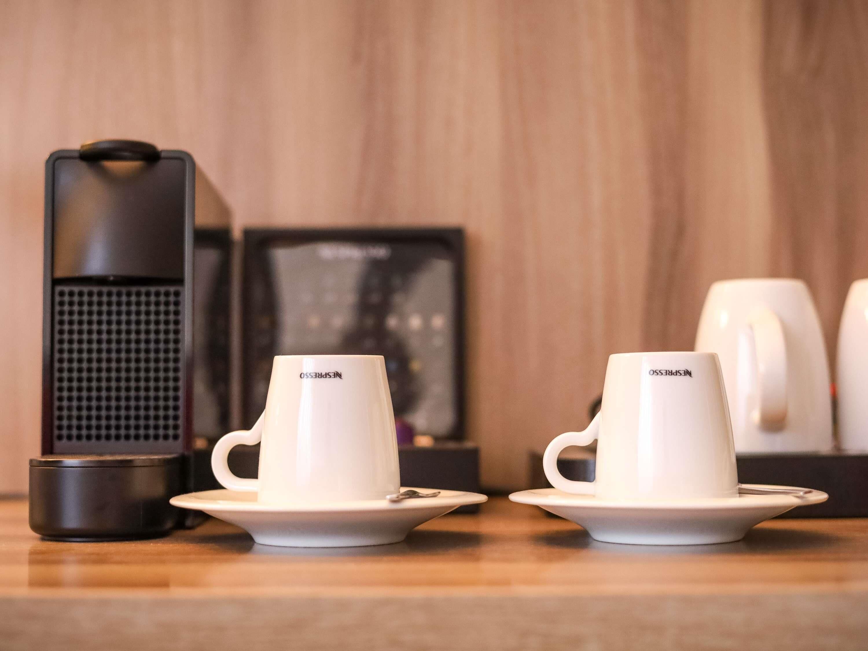 Duas chávenas brancas Nespresso em pires, uma máquina de café Nespresso e um quadro emoldurado sobre uma mesa de madeira.