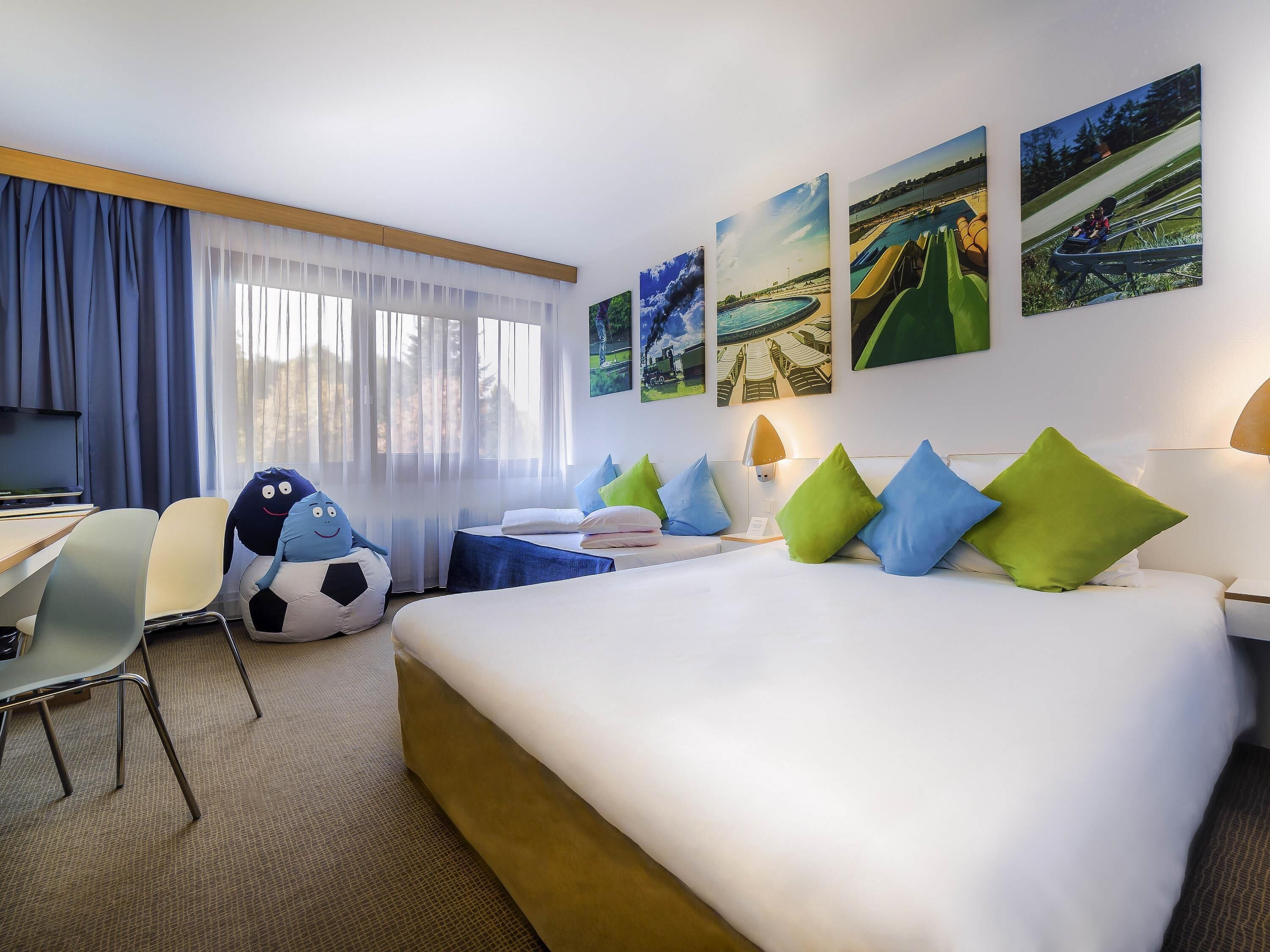 Habitación de hotel con cama, escritorio, silla y ventana con cortinas translúcidas.