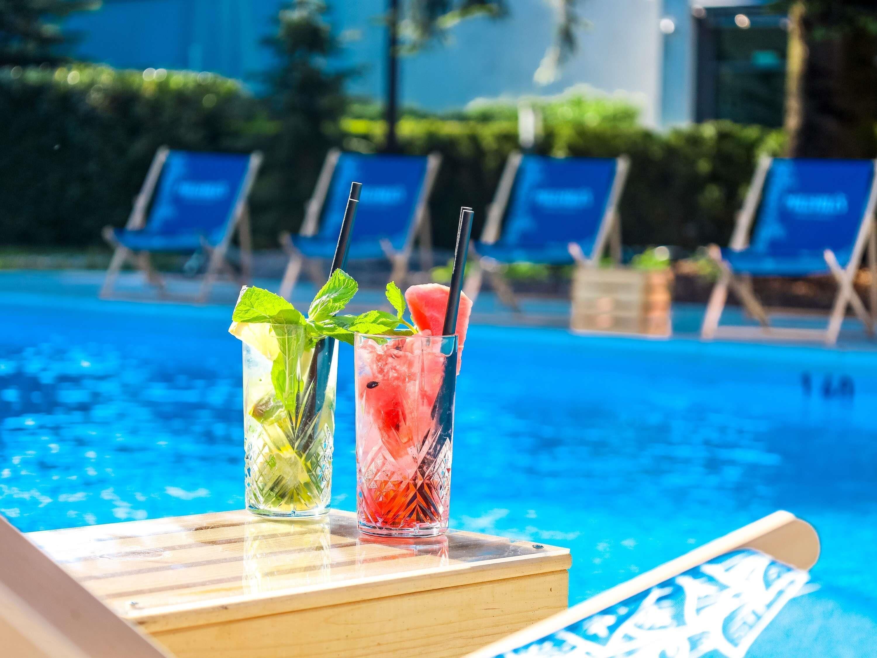 Dos bebidas sobre una mesa de madera junto a una piscina con sillones de descanso al fondo.