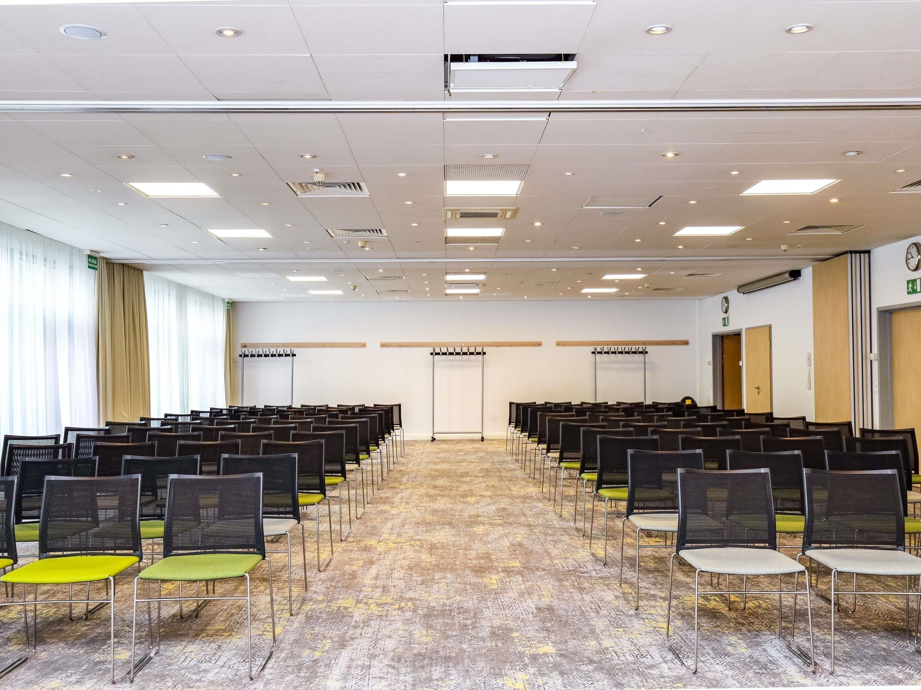 Un salón de conferencias con filas de sillas, piso alfombrado, luces en el techo y un reloj en la pared.