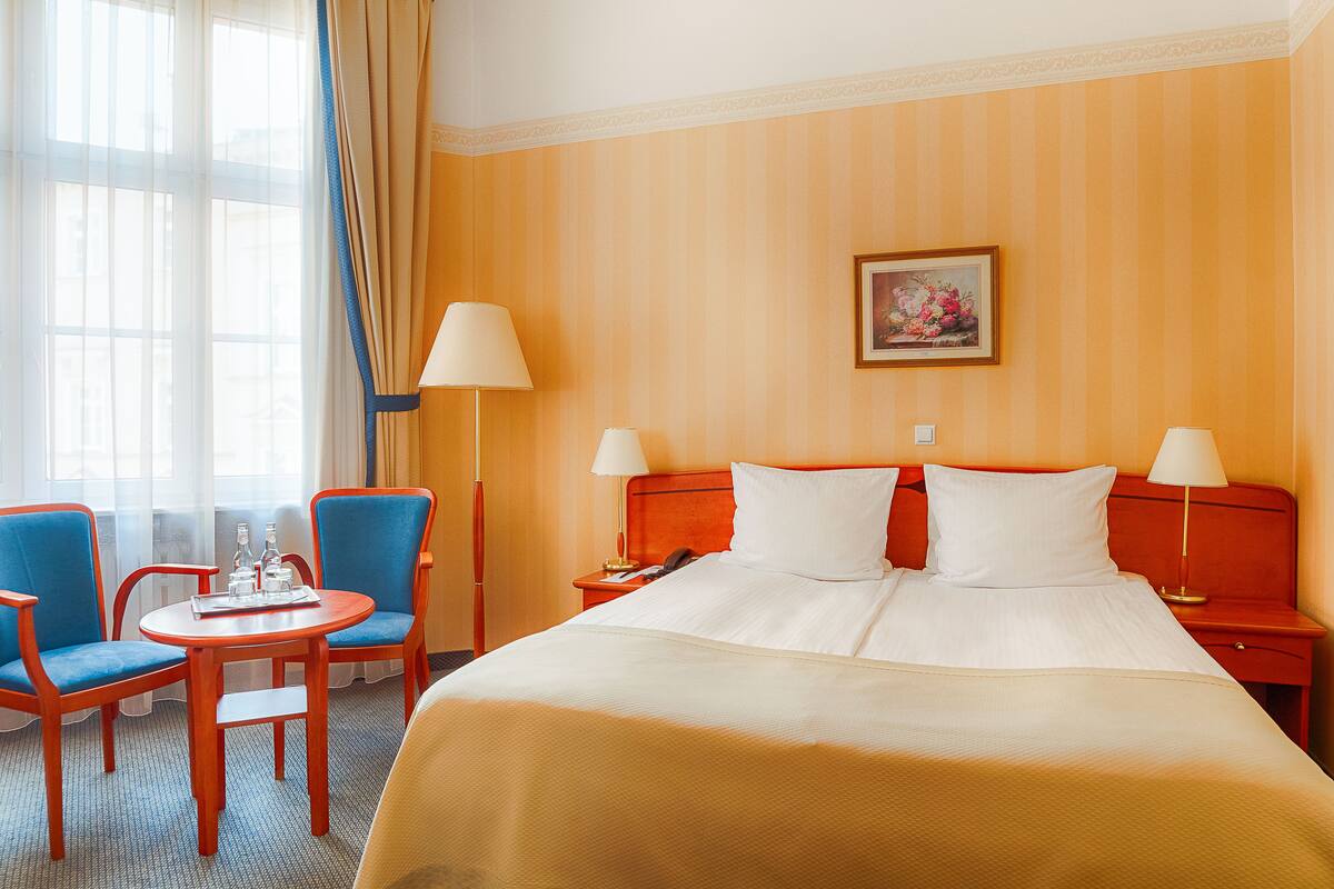 Ein Hotelzimmer mit einem großen Bett, zwei Nachttischen, einem Beistelltisch, einem runden Tisch mit zwei Stühlen und einem Fenster mit transparenten Vorhängen.