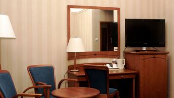 A hotel room with a wooden desk, two blue chairs, a television, a lamp, and a cabinet.
