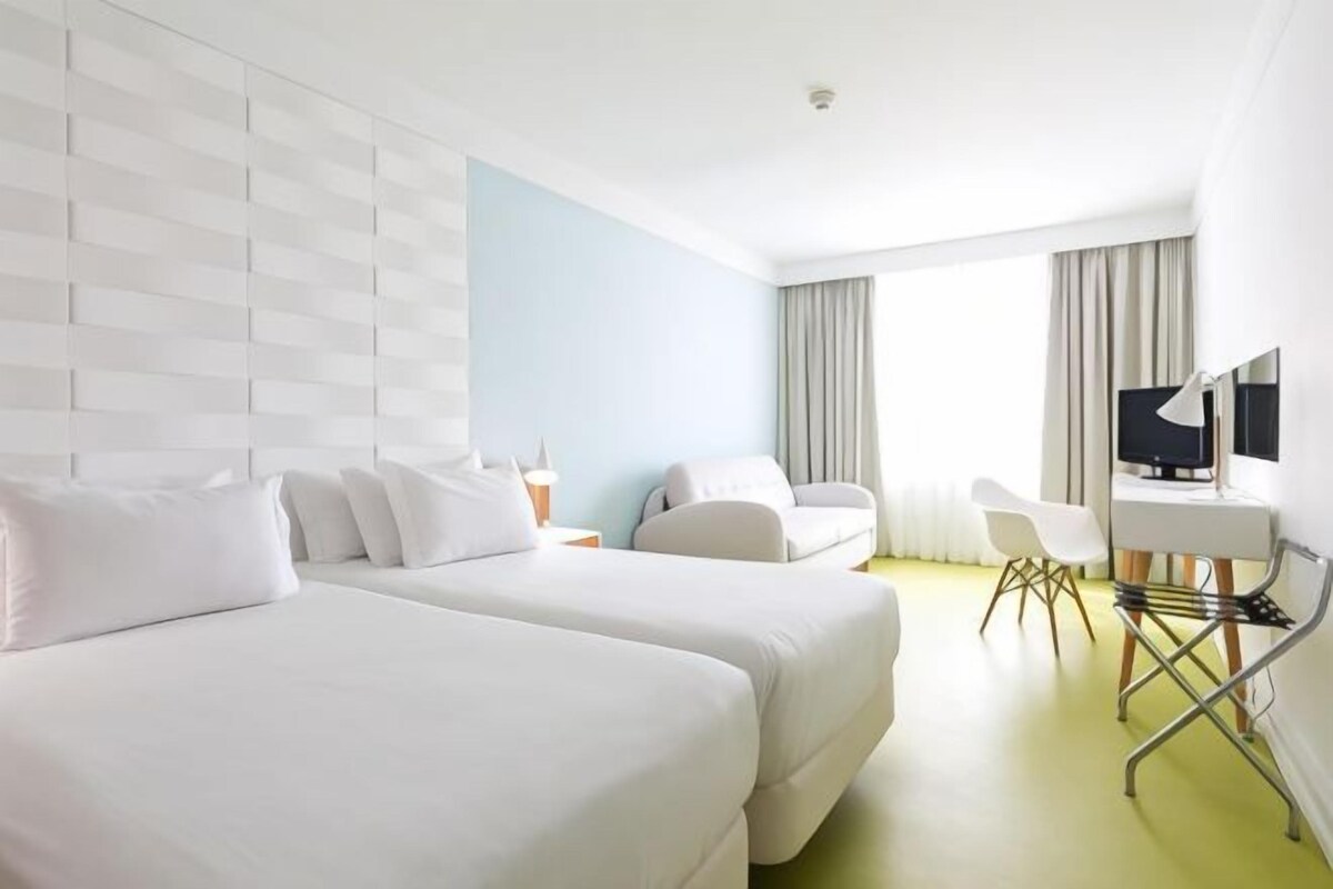 A hotel room with two beds, a desk, a chair, a TV, and a window with curtains.