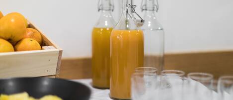 Een houten kist met sinaasappels, twee glazen flessen met oranje vloeistof, een zwarte schaal met ananasstukjes en verschillende lege glazen op een tafel.