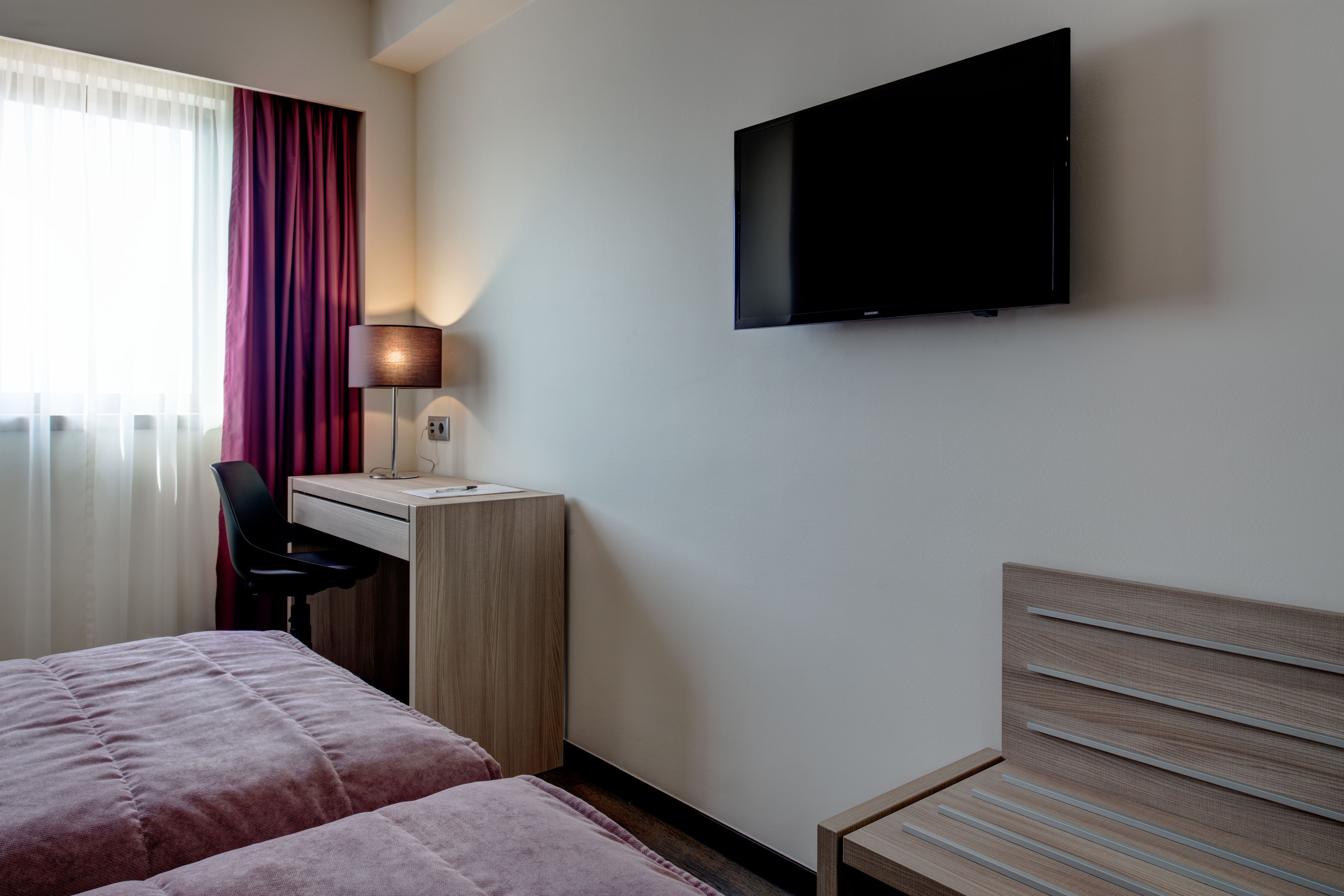 A hotel room with two beds, a desk, a chair, a TV, and a lamp.