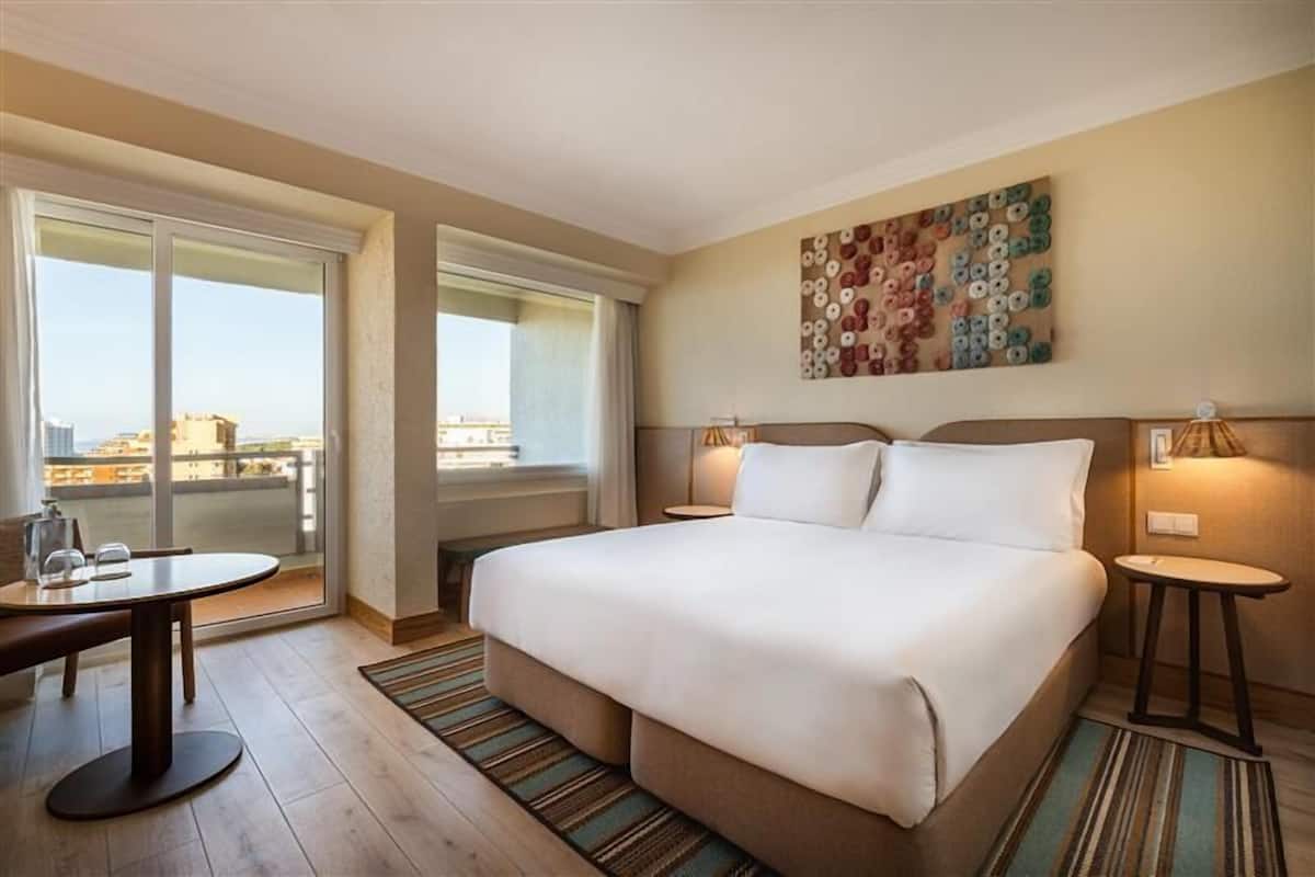 Una habitación de hotel con una cama grande, dos mesitas de noche, una pequeña mesa redonda, una ventana con vistas y una obra de arte en la pared.