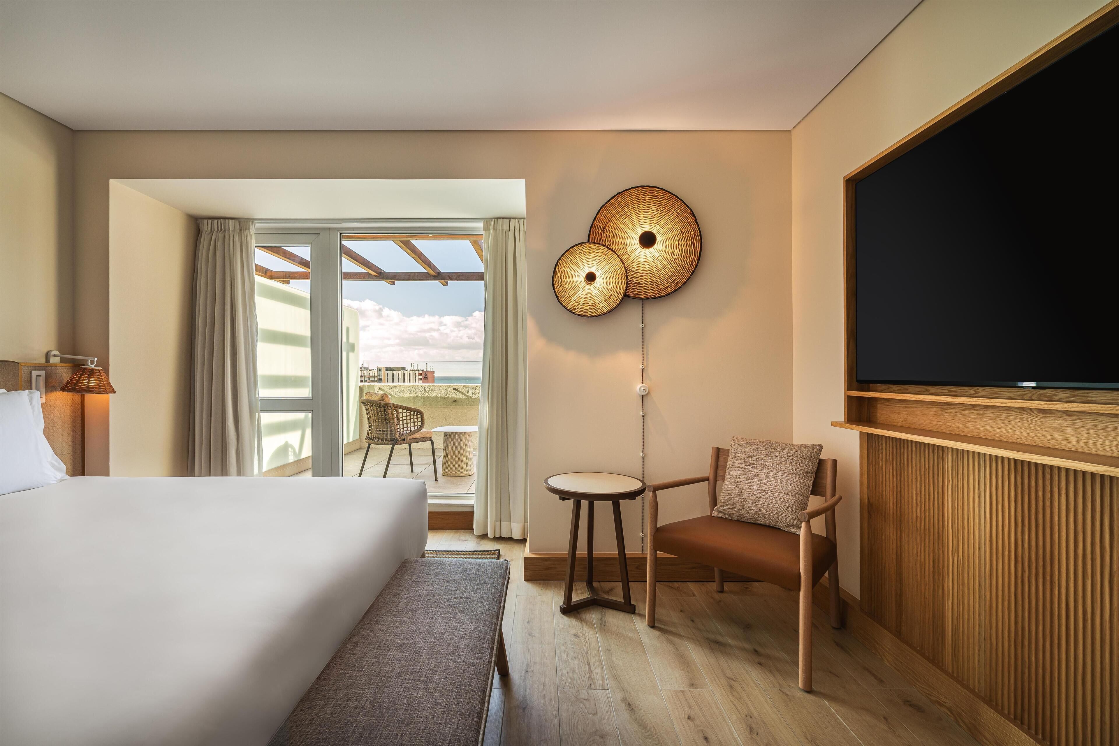 A hotel room with a bed, a TV, a small table, a chair, and a view of the outdoors.