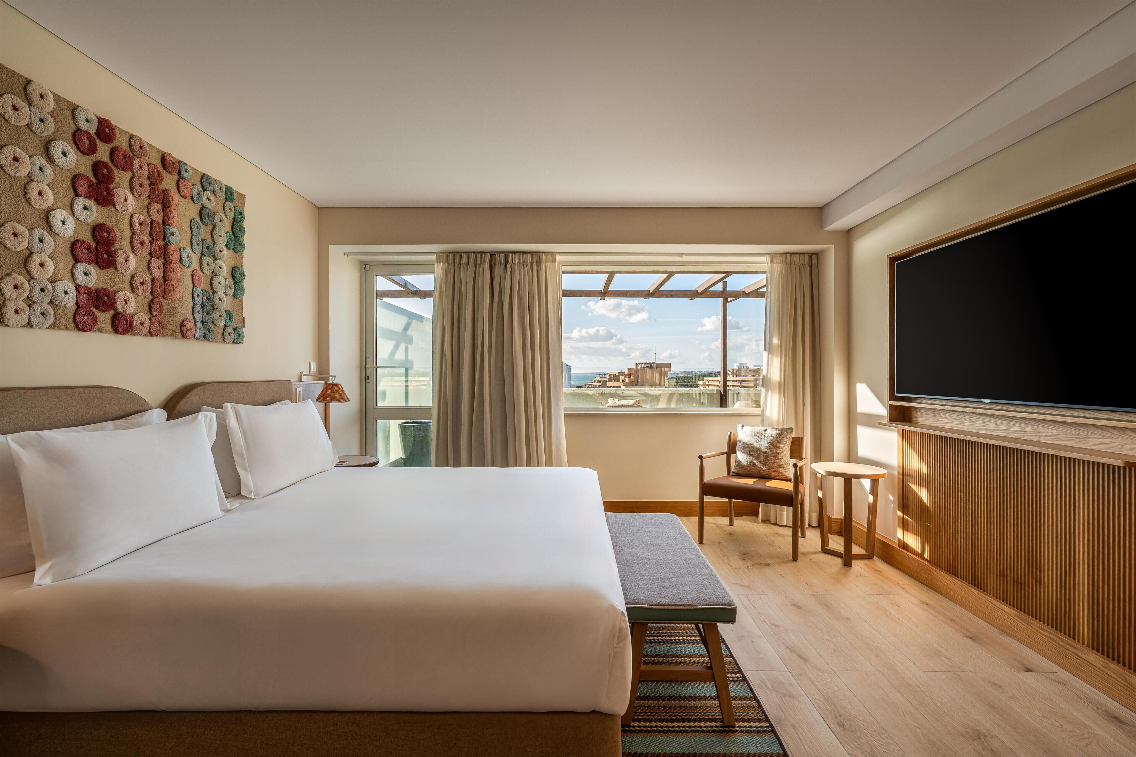 A hotel room with a large bed, a TV, a window with curtains, a chair, and a view of the city.