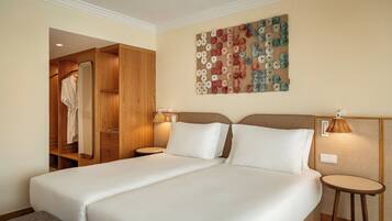 A hotel room with a large bed, wooden wardrobe, a mirror, two bedside tables with lamps, and a wall art piece.