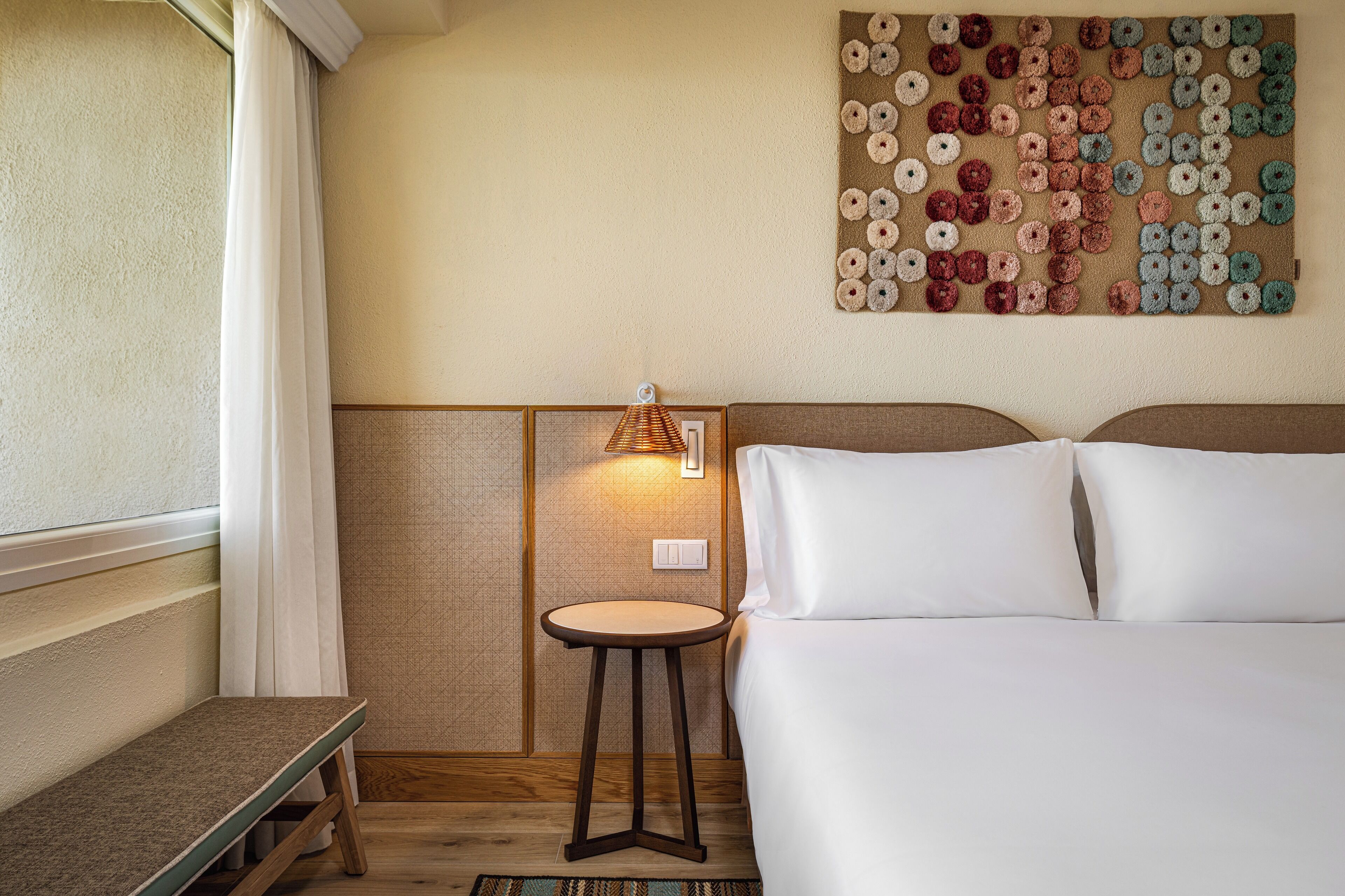 A hotel room with a bed, a small table, a wall-mounted lamp, and a decorative wall piece.