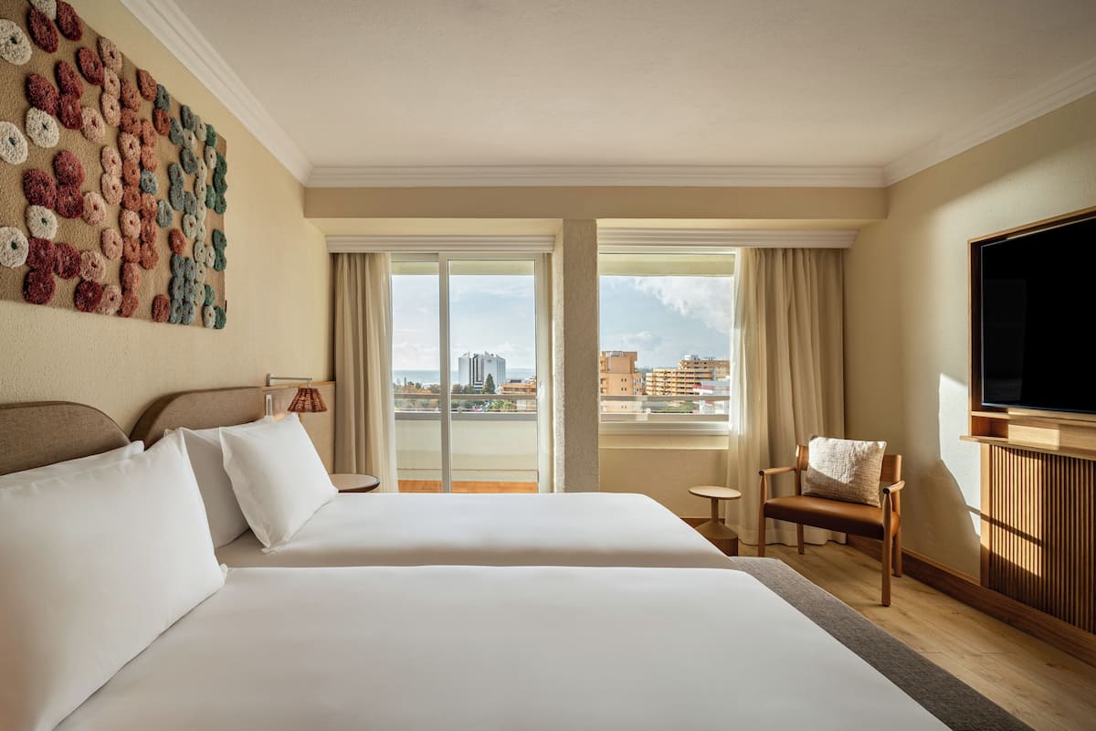 Habitación de hotel con dos camas, un ventanal grande, televisión y vista a la ciudad.