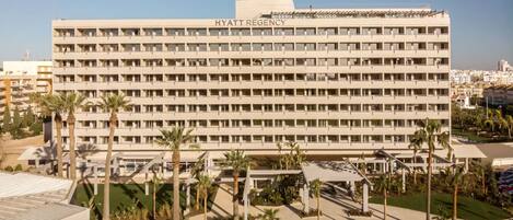 Un immeuble à plusieurs étages, sur lequel on peut lire l’inscription « HYATT REGENCY ». Il a un toit plat, de nombreux balcons et est entouré de palmiers et de verdure.