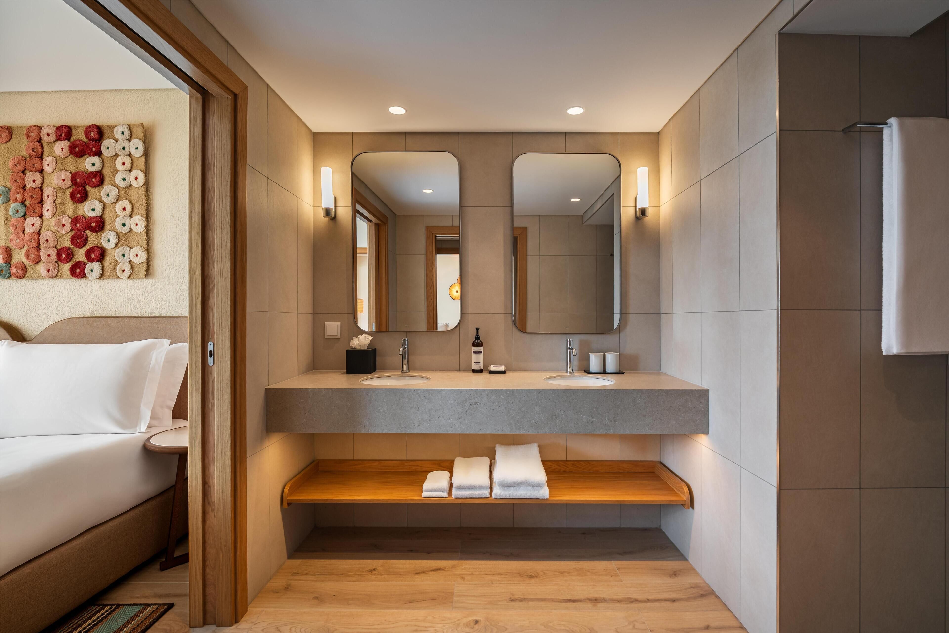 A modern bathroom with a large mirror, two sinks, a wooden shelf with towels, and a bed in the adjacent room.